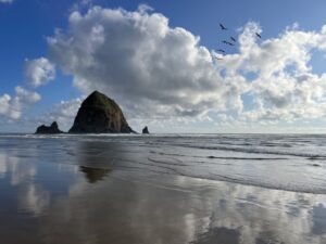 Haystack Rock in Cannon Beach, Oregon. There are clouds but the sun is out and the sky is blue. A flock of seagulls is visible.